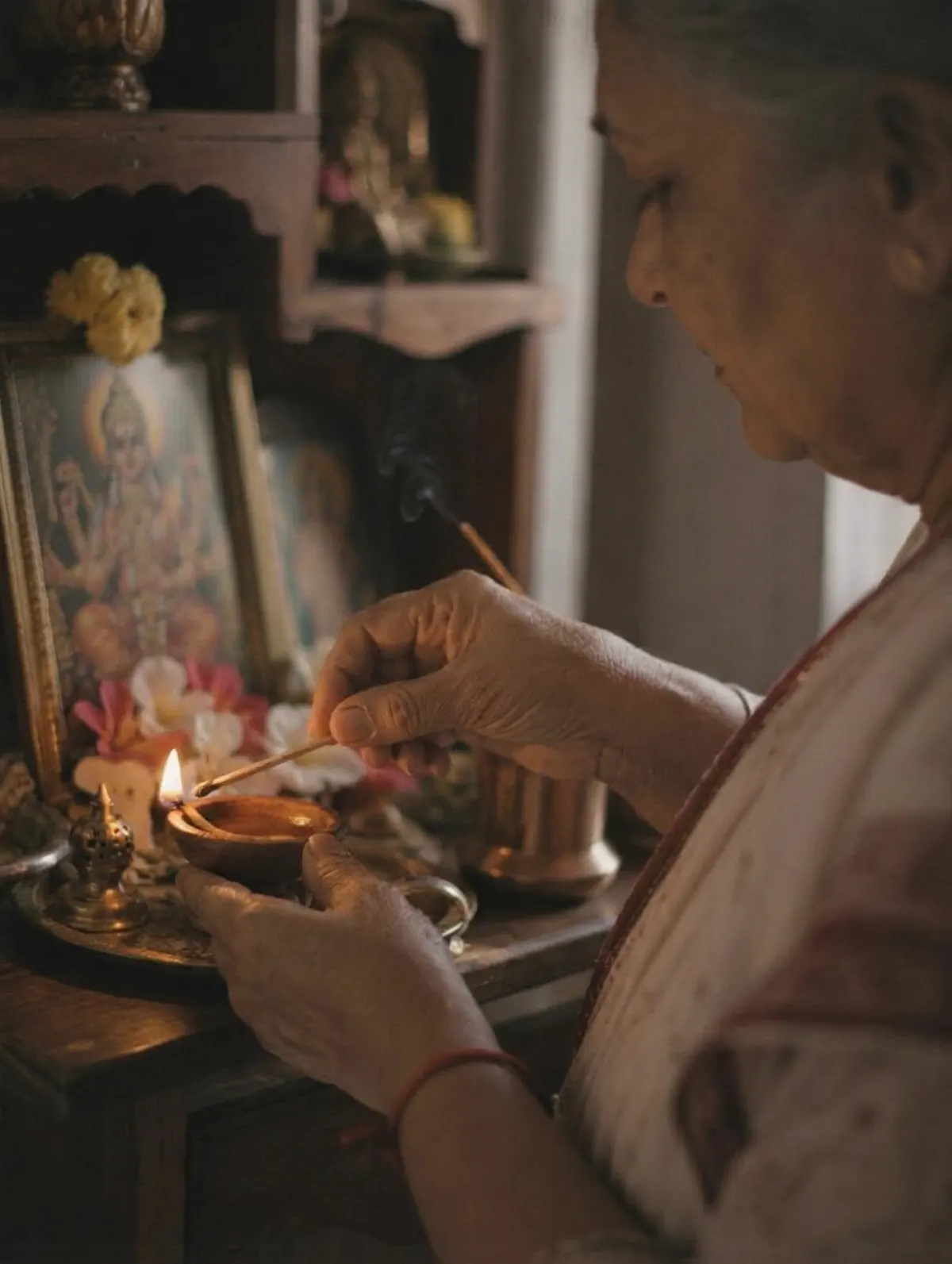 An elderly Indian woman lighting a clay diya at her home altar, with marigolds and a deity portrait in soft warm light